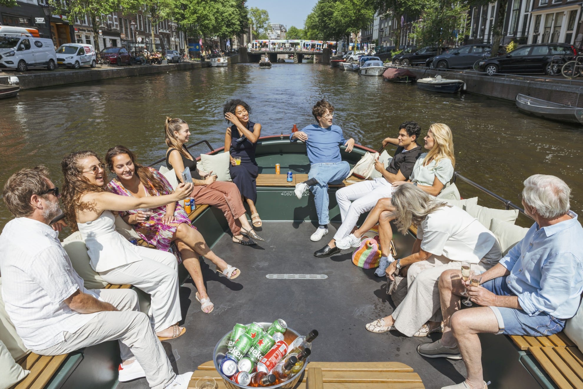 Een groep mensen geniet van een zonnige rondvaart door de Amsterdamse grachten, terwijl ze ontspannen kletsen, drinken en selfies maken op de boot.