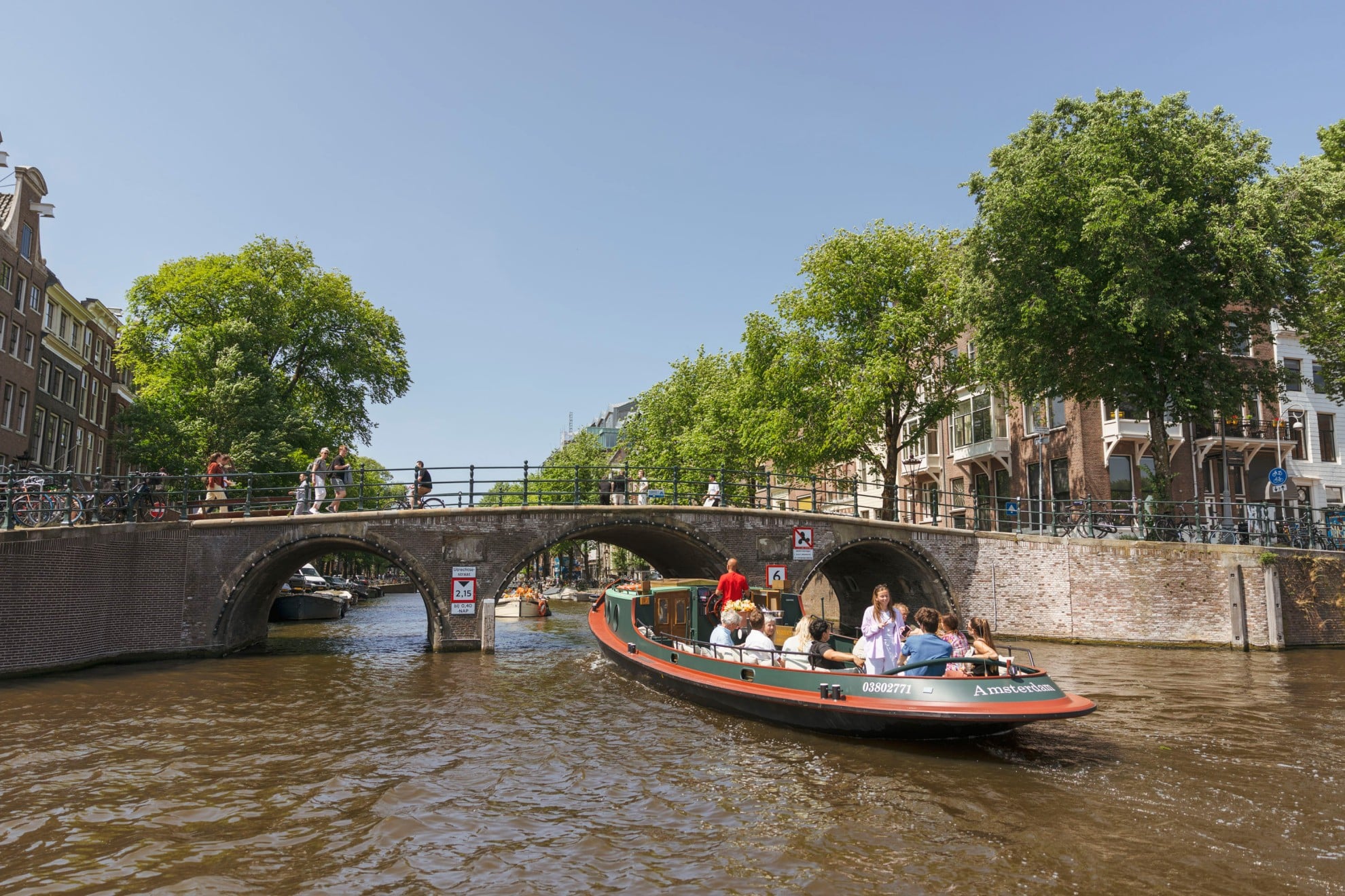 Een rondvaartboot met passagiers vaart onder een stenen boogbrug door op de Amsterdamse grachten, terwijl voetgangers over de brug lopen.